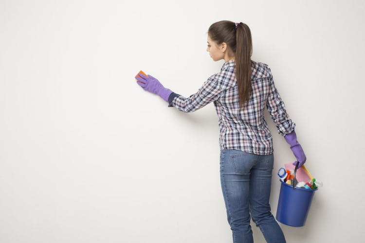La mujer limpia la pared blanca y sostiene un balde con productos de limpieza