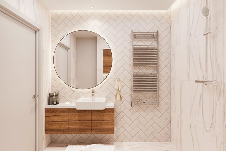 Un baño con gran espejo redondo.  El inodoro está sobre una estructura de madera y la pared tiene azulejos rectangulares.