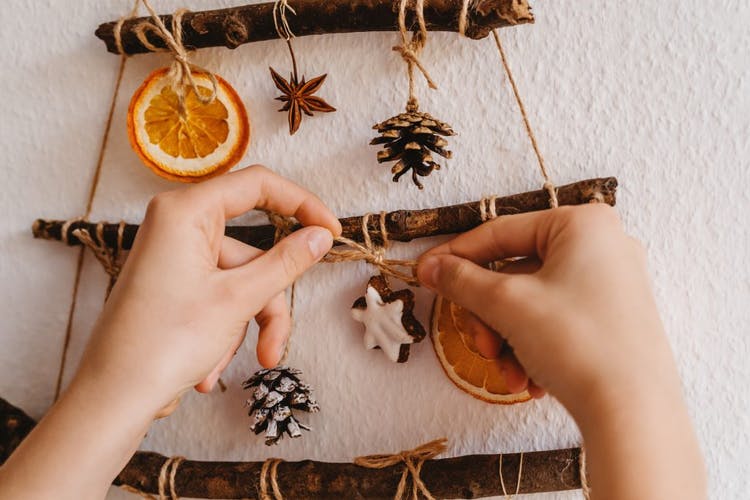 las manos de la mujer anudan cuerdas que están atadas a ramitas.  Contienen frutos secos como naranjas y pinos que forman parte de la decoración navideña.