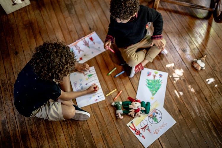 Dos niños pintando y dibujando en hojas blancas de papel.  Están sentados en un piso de madera.  Los dibujos son navideños. 
