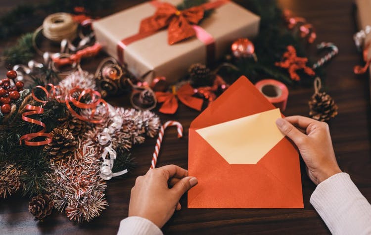 las manos de la mujer sostienen un sobre naranja con una carta de Navidad.  En la mesa de enfrente se puede ver un regalo envuelto con papel y lazo y otros adornos navideños.