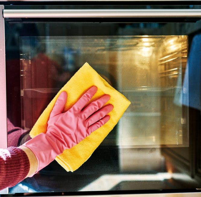 Una mujer, con un guante de limpieza rosa, limpia la puerta de un horno con un paño amarillo