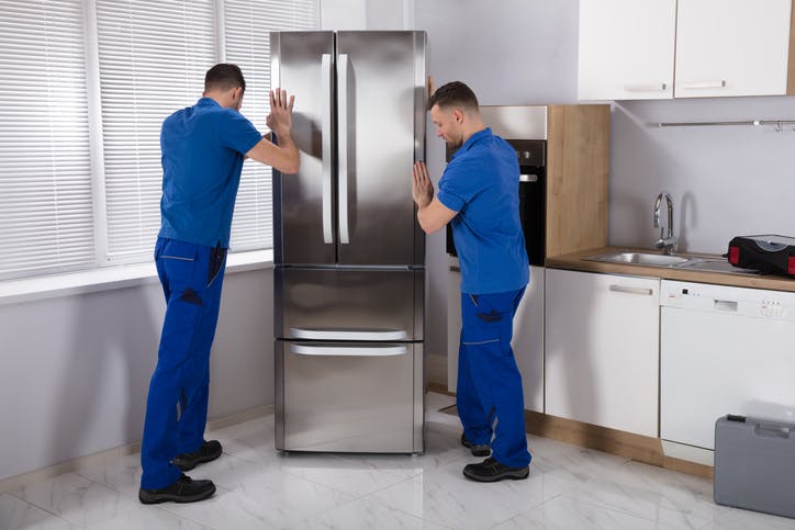 Dos hombres están empujando un refrigerador en la cocina.  Visten uniformes azules.