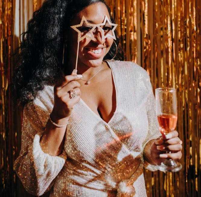 Una mujer negra está en una fiesta con un vestido de fiesta.  Ella está sonriendo y aparentemente posando para una foto mientras sostiene anteojos en forma de estrella sobre sus ojos.