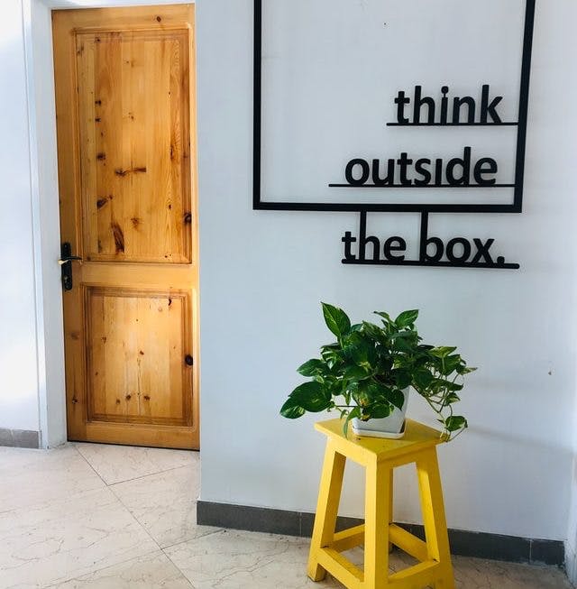 El hall de entrada de una casa.  Hay un taburete de madera amarillo con un jarrón encima.  En la pared está escrita la frase: Think outside the box