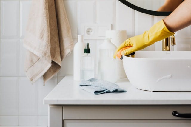Una persona está limpiando el lavabo de un baño.  Ella usa guantes de limpieza amarillos y pasa una esponja vegetal suave sobre el material.
