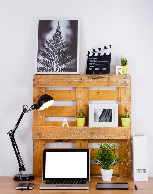 Las paletas hacen una pequeña librería sobre un escritorio en casa.  Es posible ver pinturas, plantas, una computadora portátil y una lámpara de mesa.