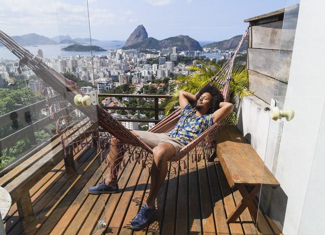 Un hombre está tumbado en una hamaca en el porche de su casa.  Está en Río de Janeiro y al fondo se ve el Monumento Natural del Pan de Azúcar.