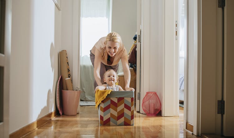 Una madre juega empujando a su bebé dentro de una caja sobre un piso de madera.  Ambos se divierten y sonríen.