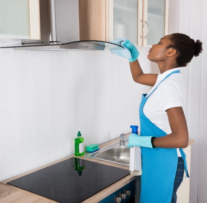 Una mujer está limpiando un extractor de aire de la cocina.  Lleva un delantal azul y está mirando por encima de los aparatos ortopédicos.