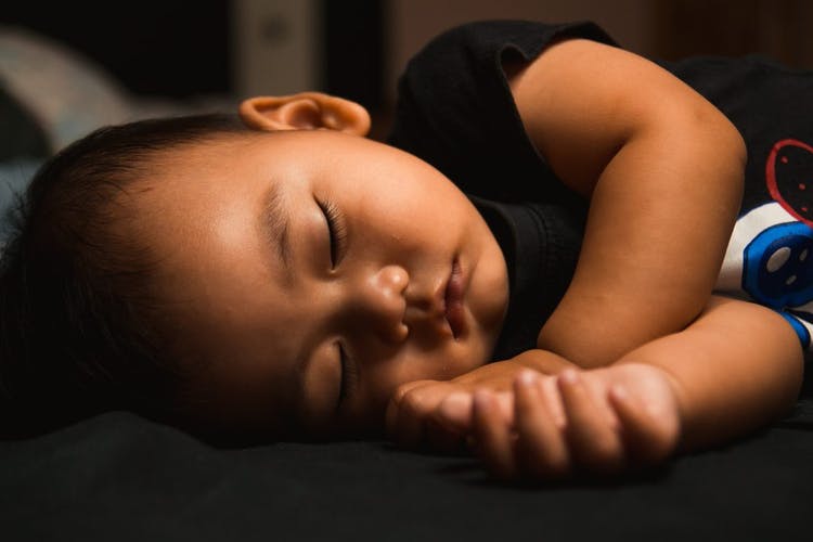 bebe durmiendo  Lleva una camiseta de color oscuro y tiene los ojos cerrados.  Solo es posible ver su torso, cara y manos.