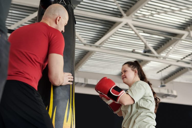 Una niña se prepara para golpear un saco de boxeo.  Lleva guantes de boxeo rojos y tiene síndrome de down.